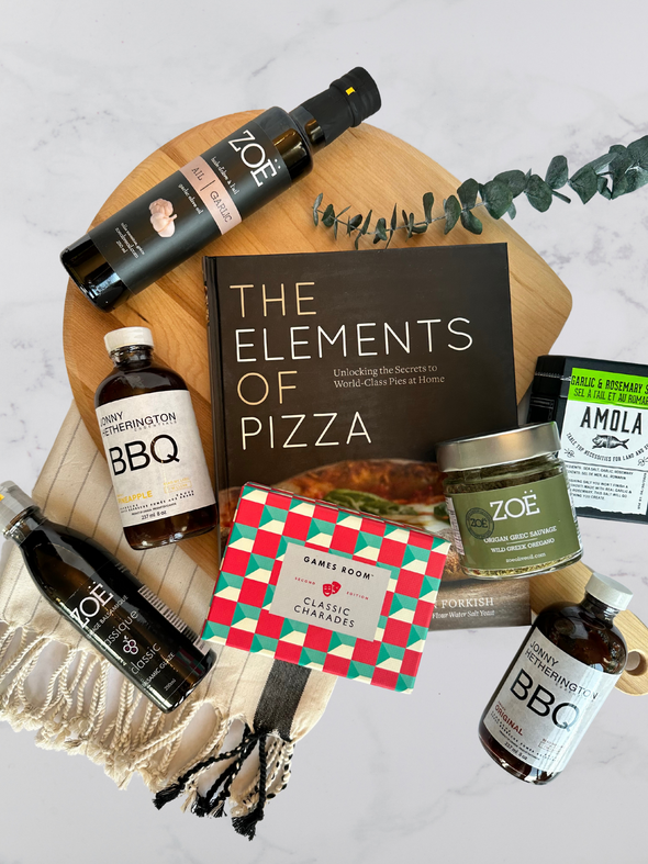 This is a picture of the contents of the Ultimate Home Pizza Experience Gift Basket. It's a flatlay of the wooden pizza paddle, flavour infused olive oil, organic bbq sauces, balsamic glaze, oregano and garlic and rosemary sea salt and the elements of pizza cookbook.